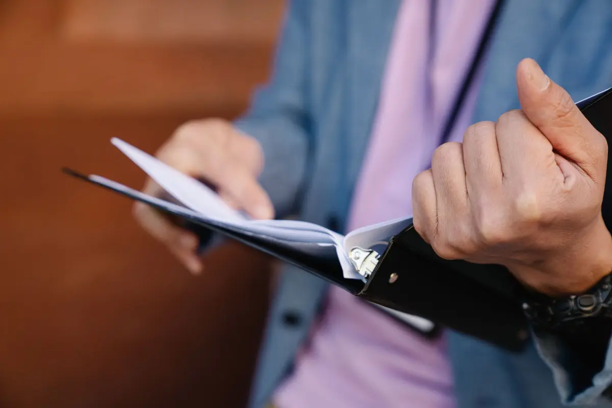 Hands flip through the pages of a black ring binder, the kind an operations manual comes in, held against a blue jacket.