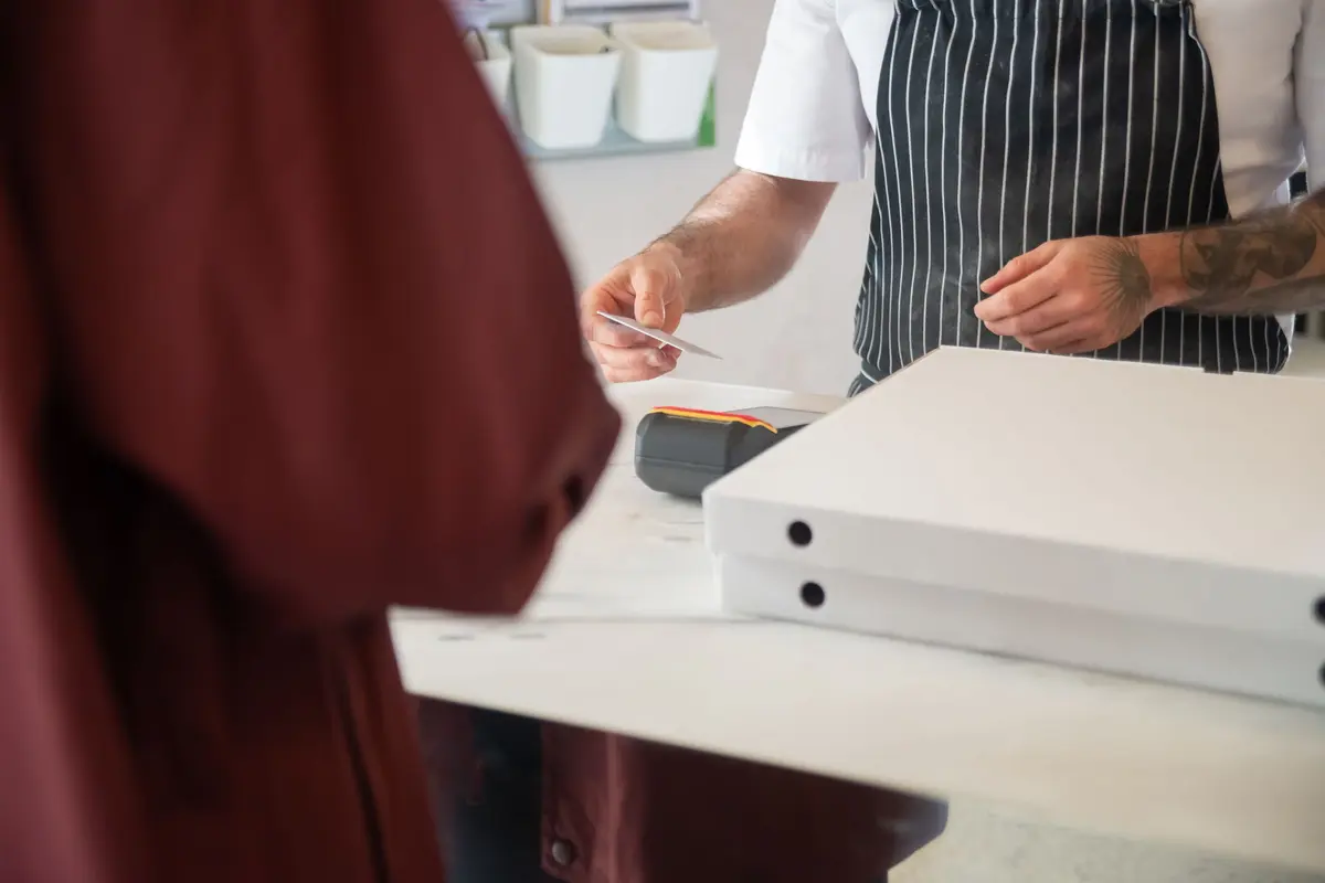 Two plain pizza boxes wait on a pickup counter while a worker takes a card payment.