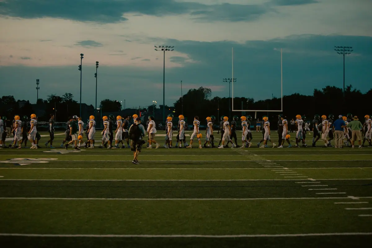 High-school football players walk across the field at dusk, small in the distance, under tall light poles.