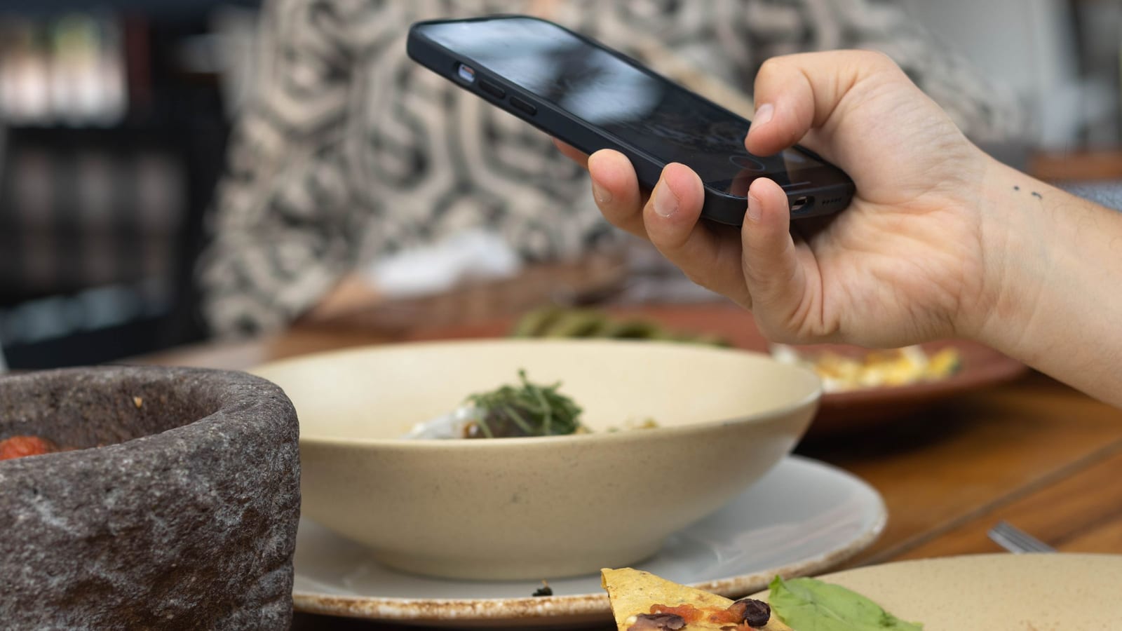 A hand holds a phone over a restaurant table to photograph a bowl of food, more plates beside it.