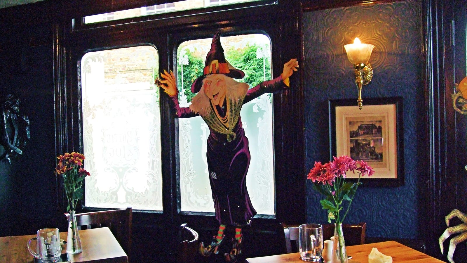 A pub dining room dressed for Halloween, with a witch cutout hanging in the window, flowers in vases and empty glasses on set wooden tables.