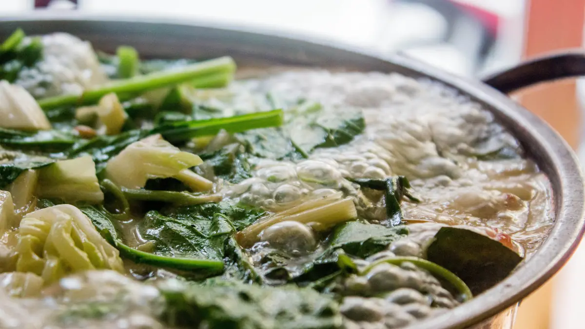 A steel hot pot at a hard boil, packed with leafy greens and cabbage.