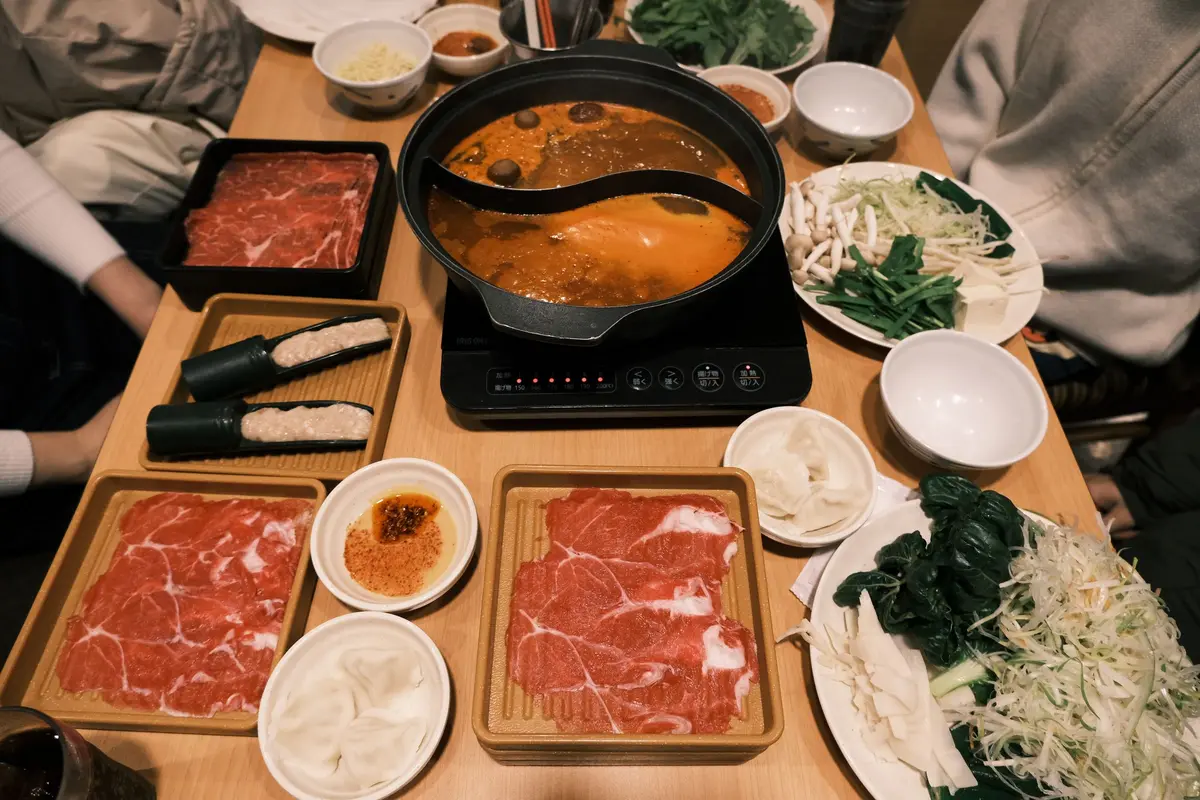 A split pot of broth on a table burner, ringed by trays of sliced beef, dumplings, greens and sauce bowls, with diners’ arms at the edges.