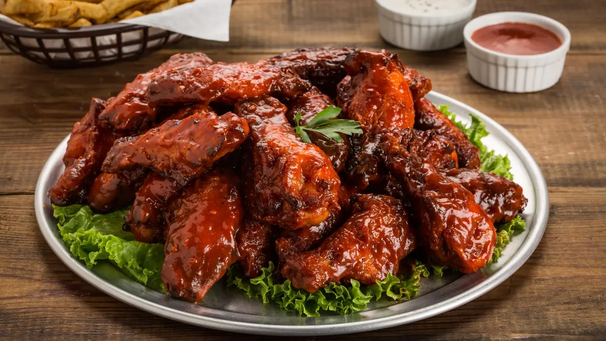 A metal platter piled with saucy barbecue chicken wings on lettuce, with two dips and a basket of fries, on a wooden table.