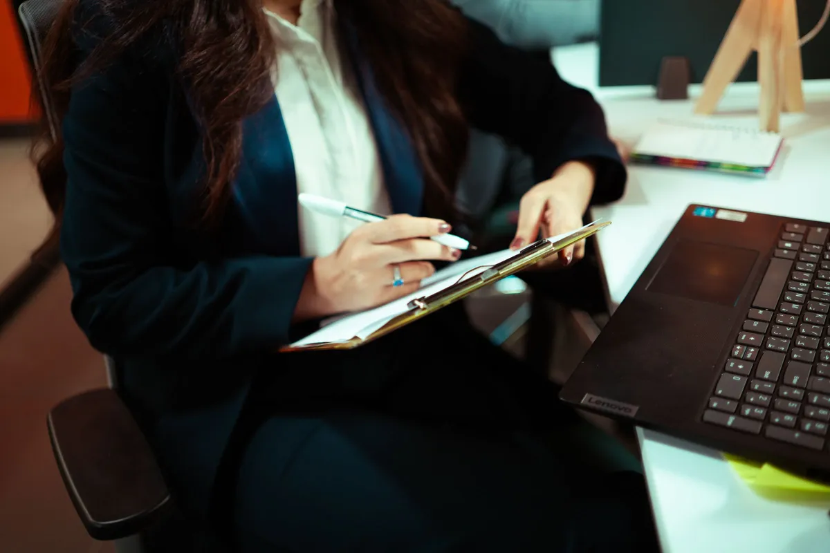 A woman in a dark blazer writes on a clipboard beside an open laptop.