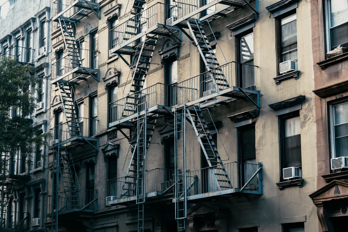 A row of brick walk-up apartment buildings with black fire escapes on a New York street.