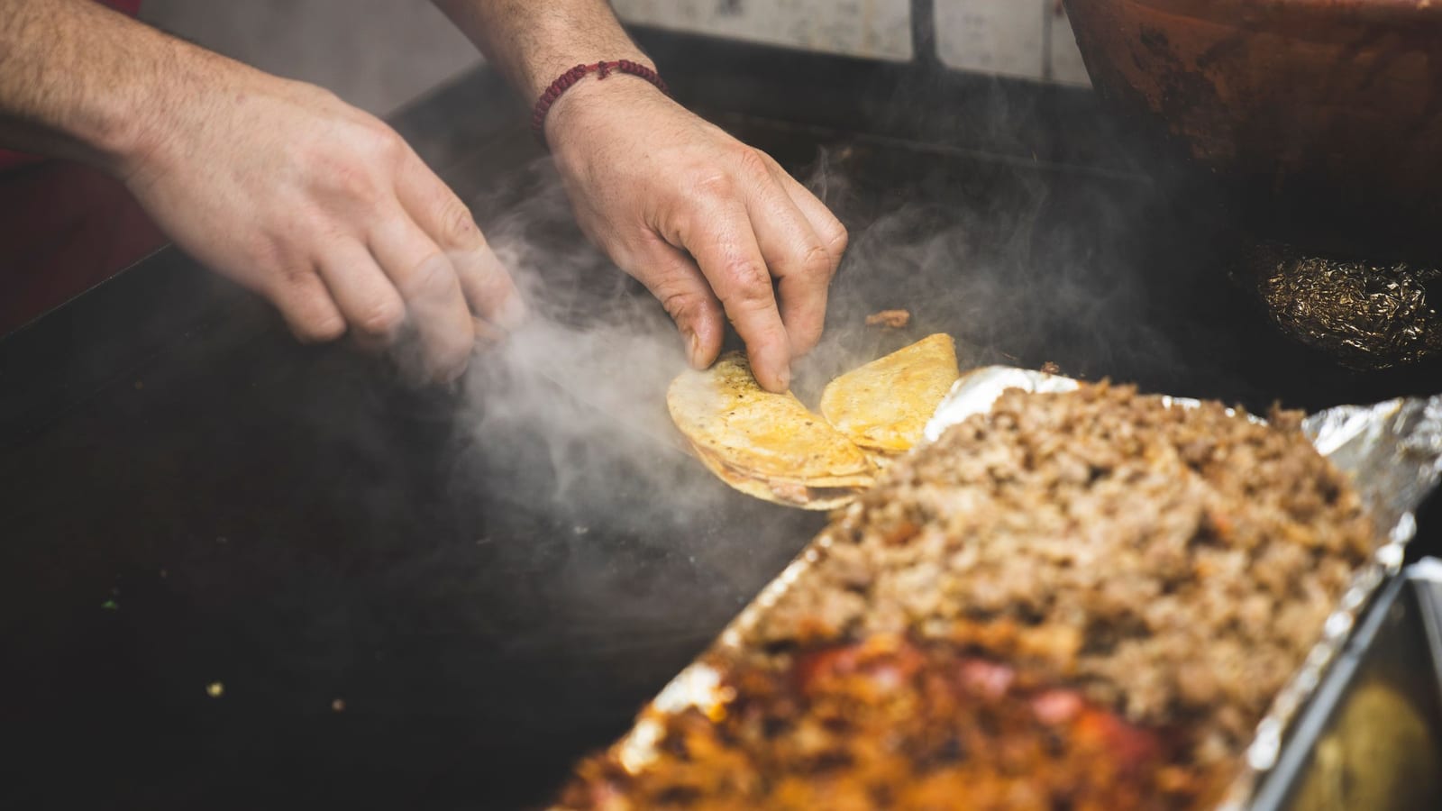 A cook’s hands fold small corn tortillas on a steaming plancha beside a tray of chopped meat.