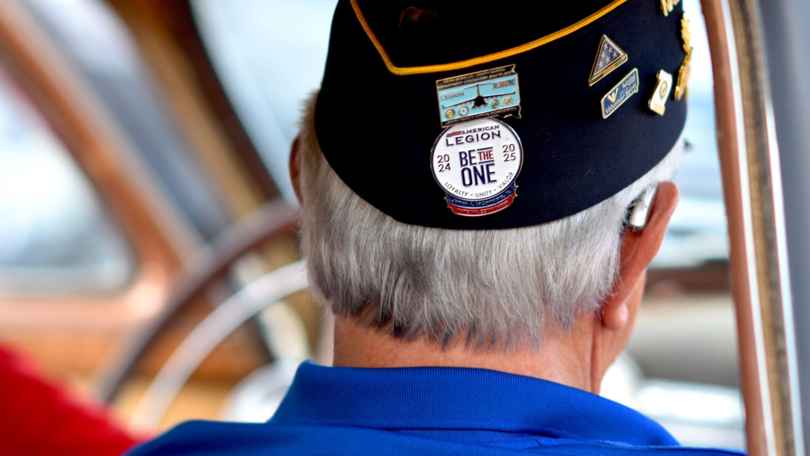 Seen from behind, a gray-haired veteran wears an American Legion cap covered in pins, the kind of cap veterans wear on Veterans Day.