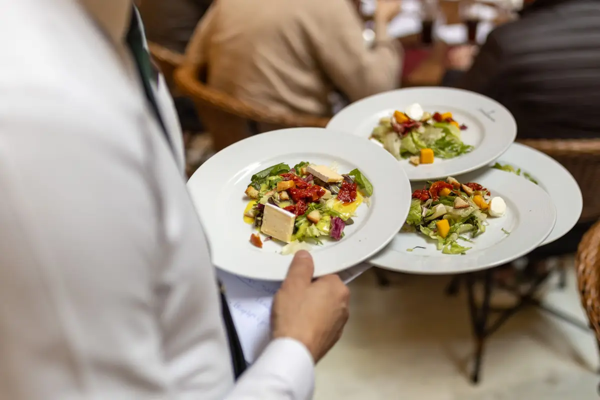 A server in a white shirt carries three plates of salad through a full dining room.