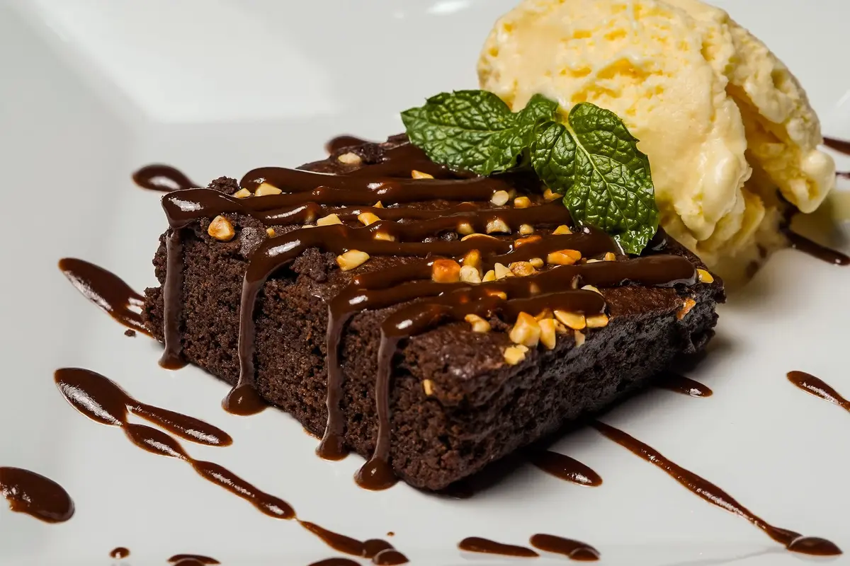 A chocolate brownie drizzled with chocolate sauce and chopped nuts sits beside a scoop of vanilla ice cream and a mint sprig.