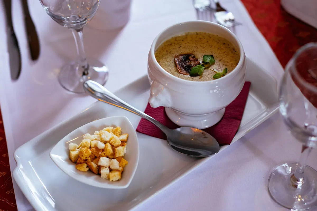 A bowl of mushroom soup with a dish of croutons on a restaurant table set with a white cloth and wine glasses.