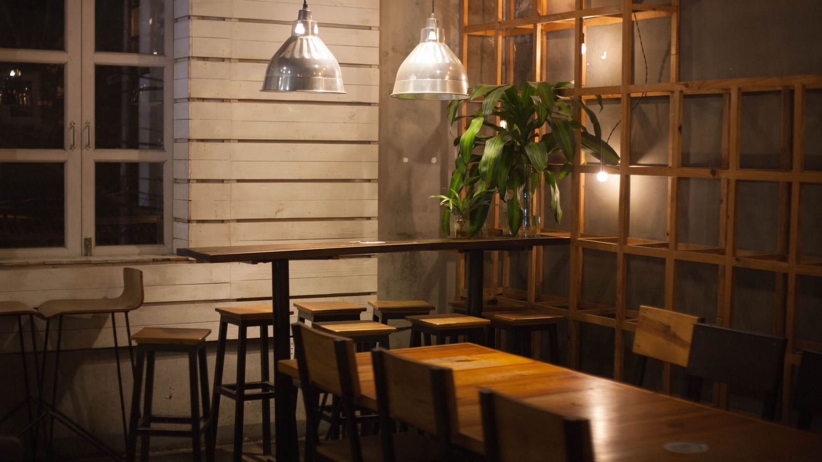 A quiet restaurant dining room in the evening, empty wooden tables and stools under two pendant lamps.