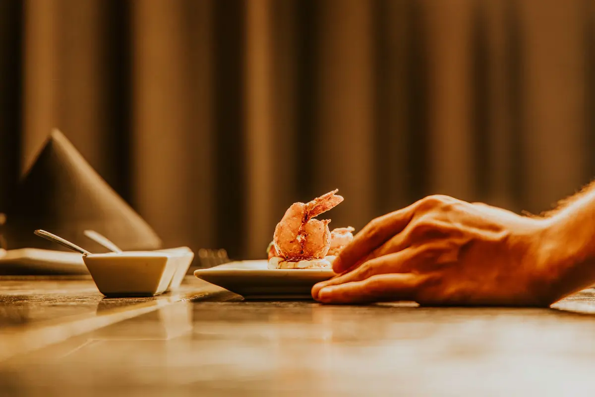 A hand rests beside a small plate of fried shrimp on a dim wooden counter.