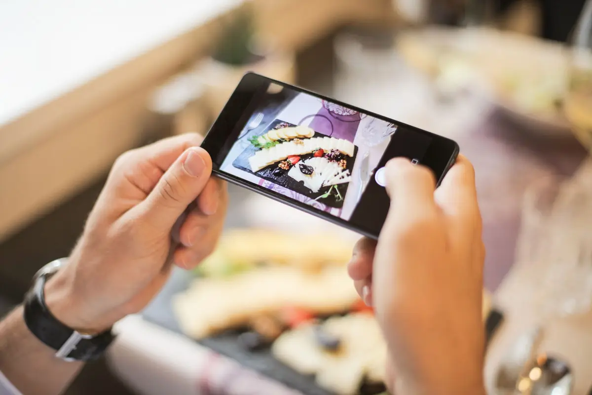 Two hands hold a phone sideways over a plate, framing the dish on the camera screen.