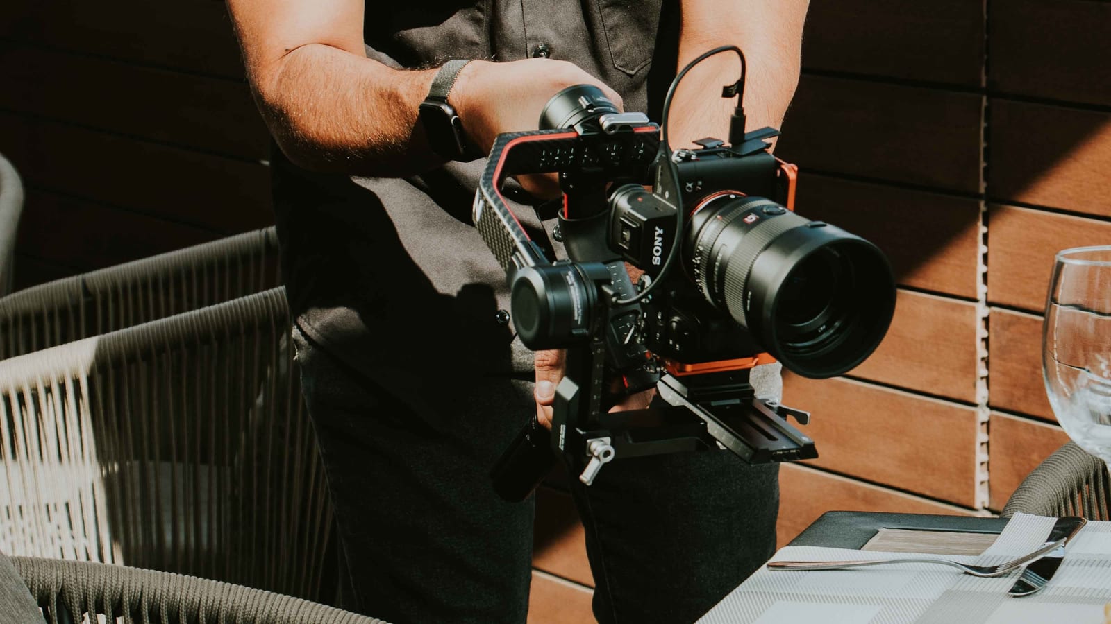 A creator steadies a camera on a handheld rig over a set table on a sunny restaurant patio.