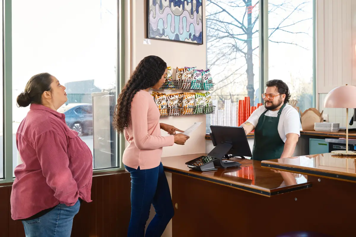 A counter worker in a green apron greets two guests at the register of a bright neighborhood restaurant.