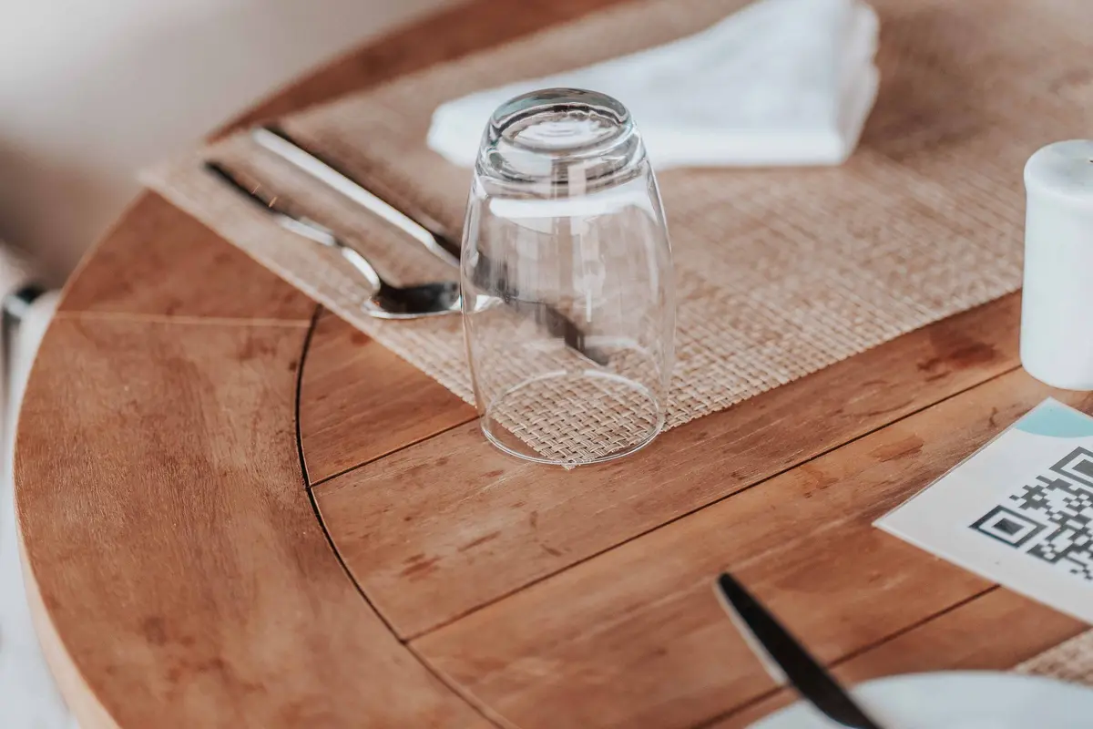 A QR code card sits on a wooden restaurant table beside an upturned glass, a napkin and cutlery.