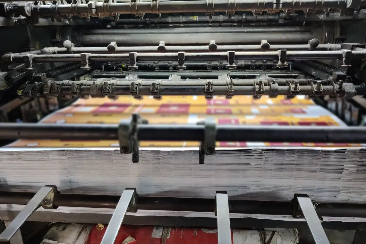 A tall stack of freshly printed sheets builds up on the delivery pile of an offset printing press, the newest sheets still under its rollers.