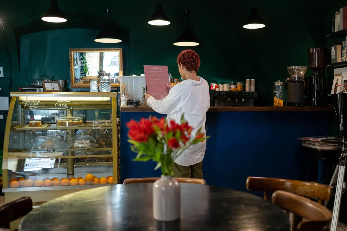 In an empty café, a woman reads a printed sheet by the counter, a pastry case beside her.