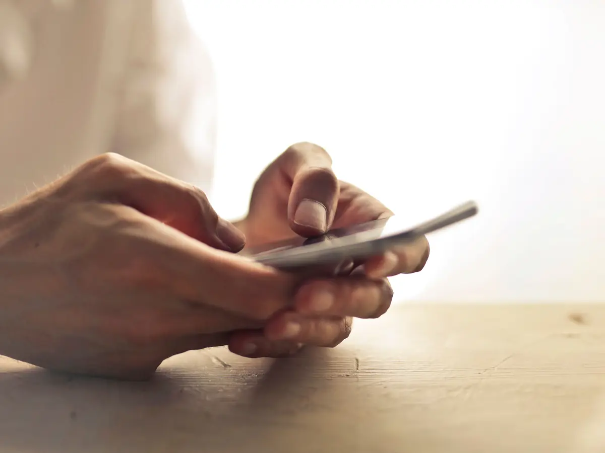 Two hands type on a smartphone held over a wooden table.