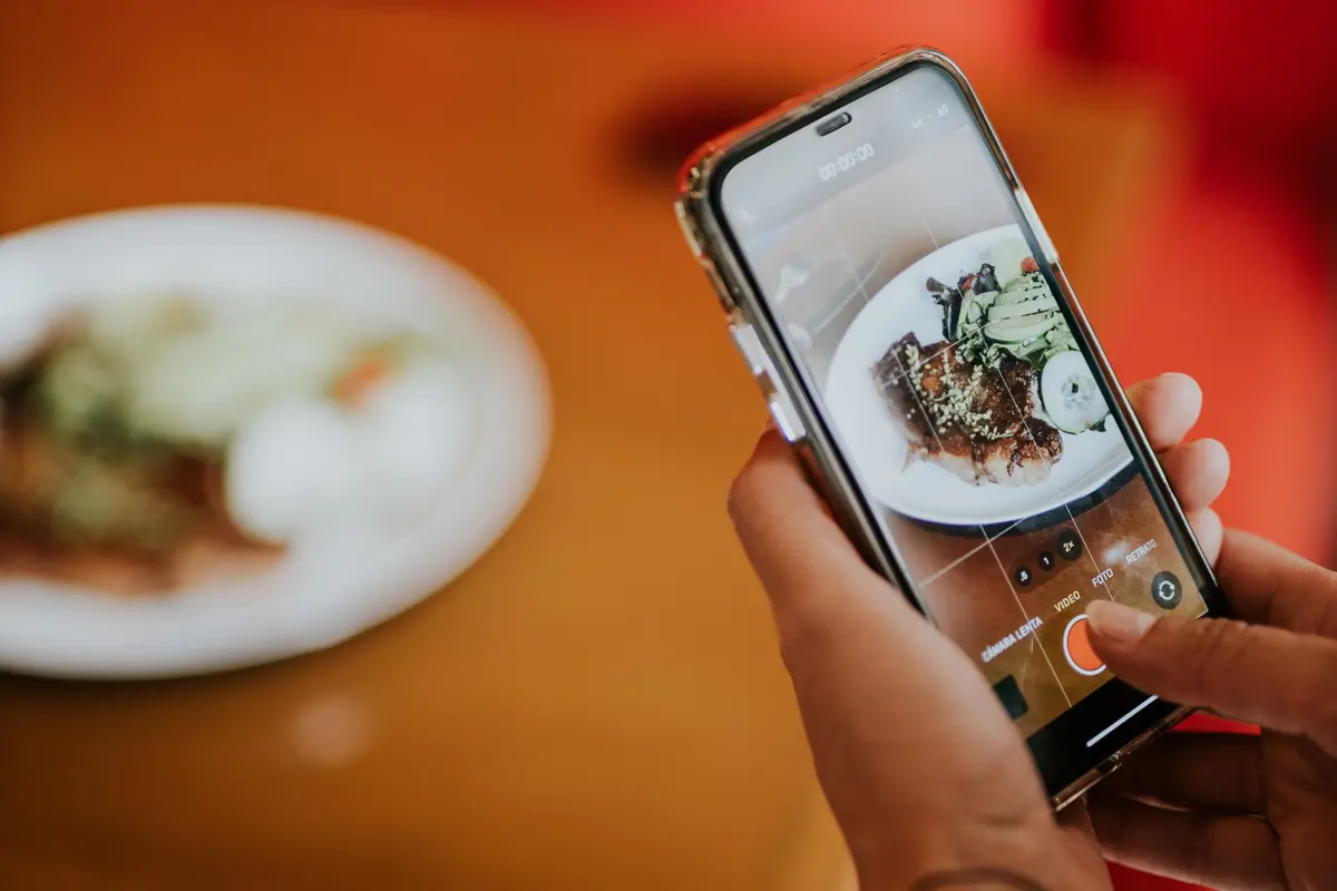 Two hands hold a phone that frames a plated dish on a restaurant table.
