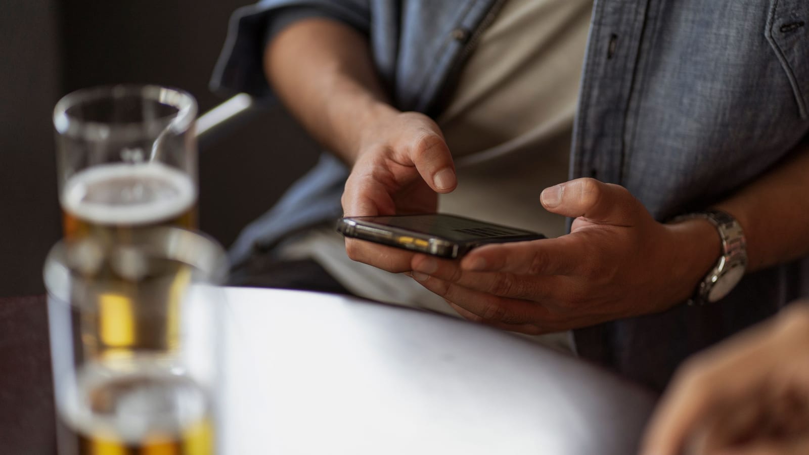 Hands type on a phone over a table, a glass of beer blurred in the foreground.