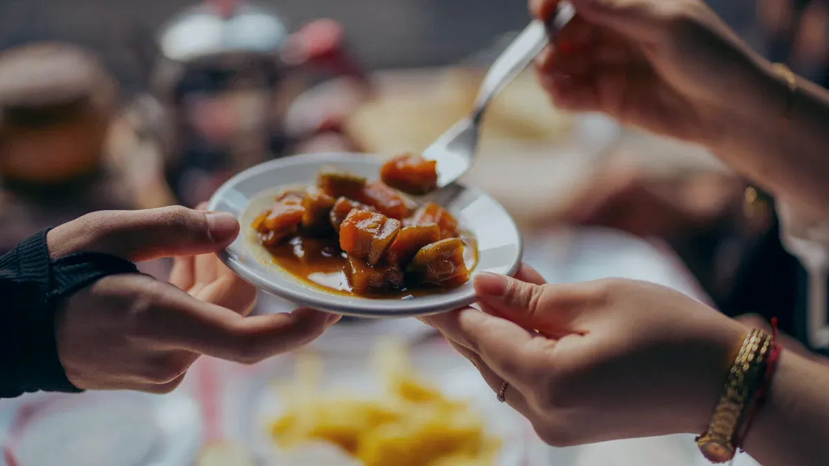 One hand passes a small plate of glazed stew across a table to another, a fork already reaching in