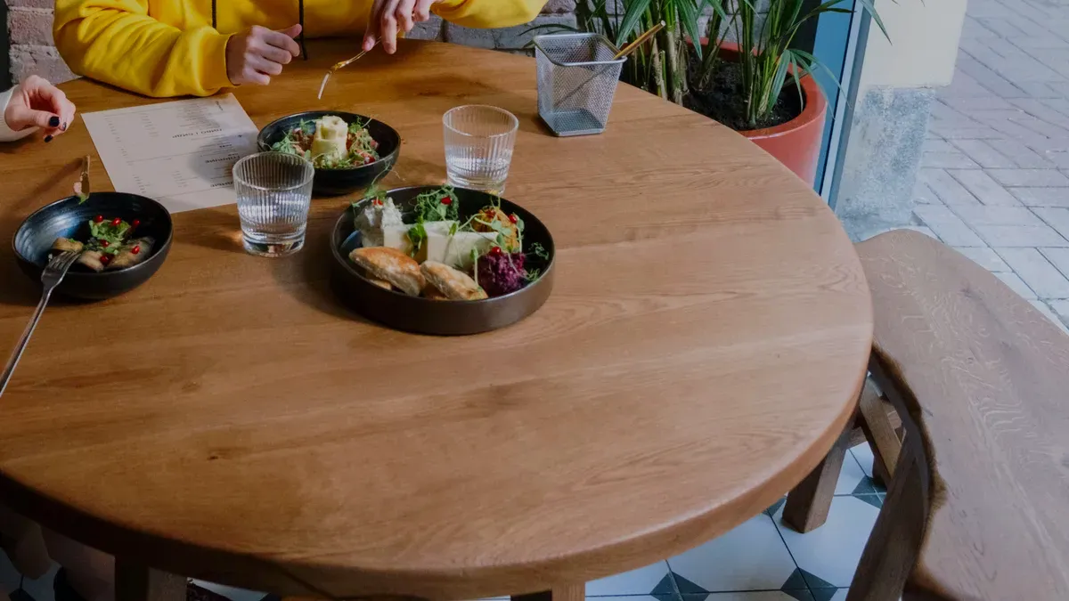 A round wooden table set with three small plates, two water glasses and a menu, two guests seen only by their hands