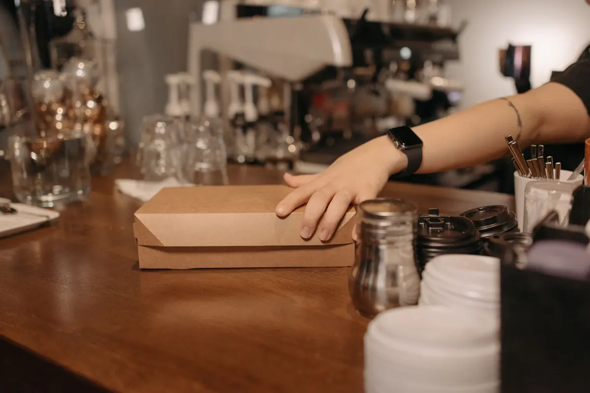 A staff member’s hand slides a kraft takeout box across a wooden café counter toward a guest.