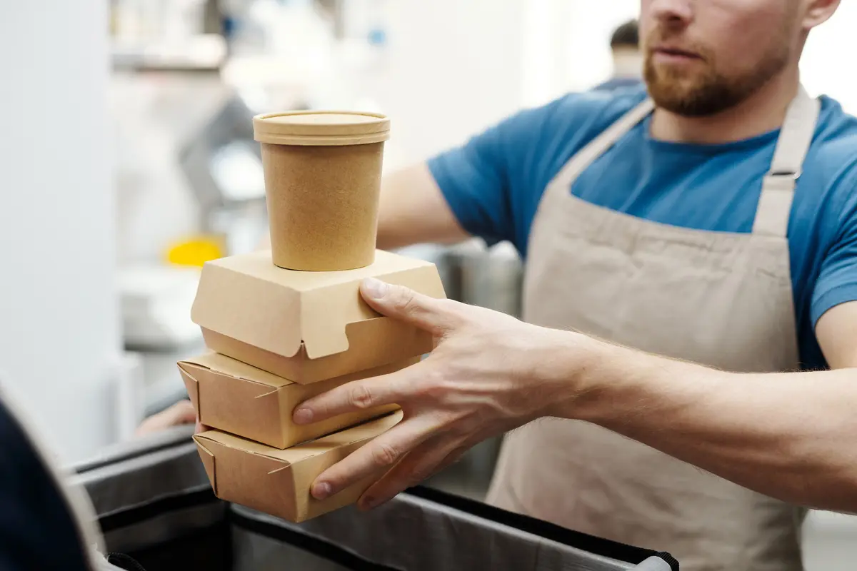 A restaurant worker in an apron lowers a stack of kraft takeout boxes and a soup cup into a delivery bag.