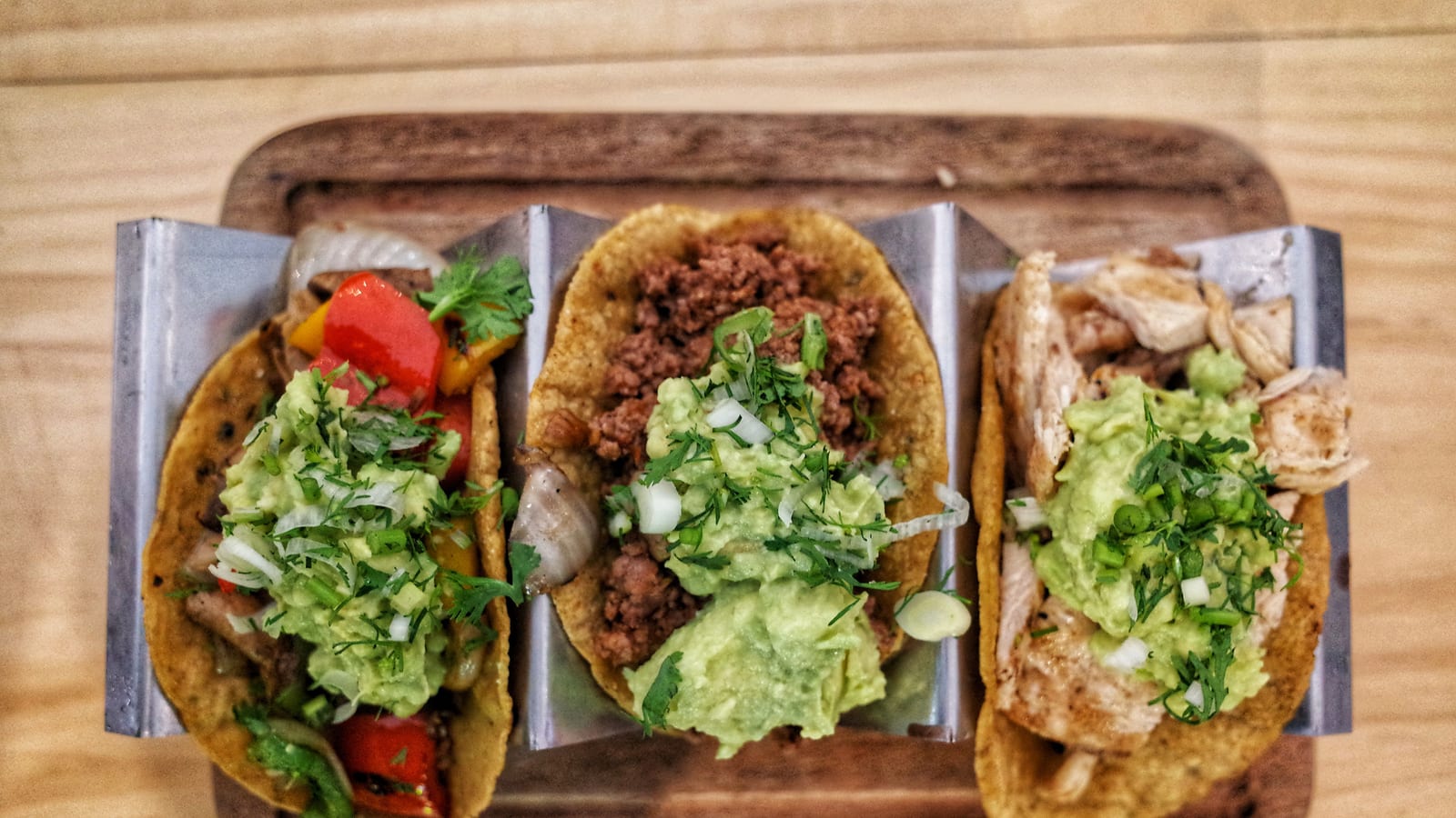 Three tacos with different fillings stand in a steel holder on a wooden tray, the kind of limited-time special that needs its own price.