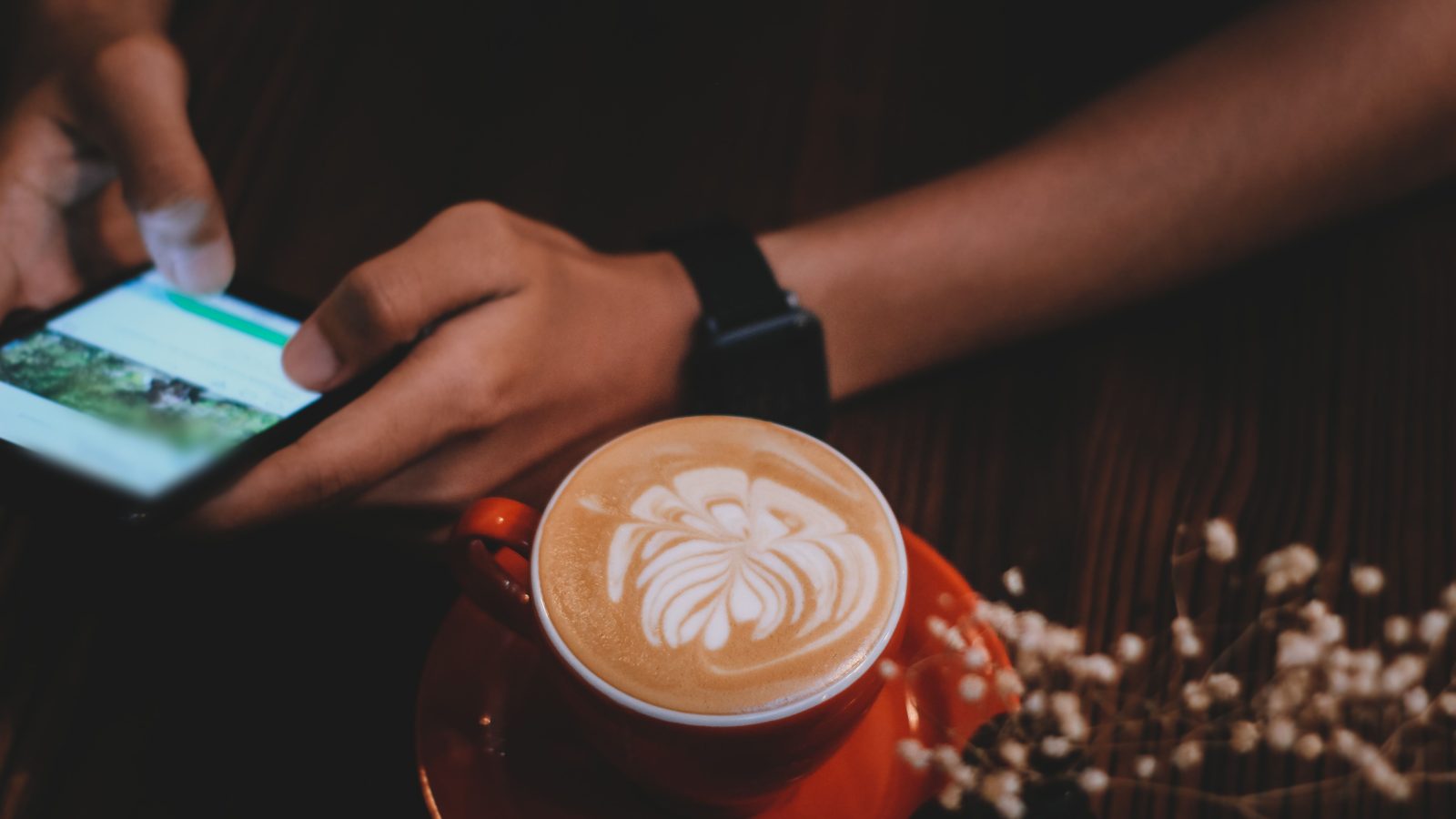 A person’s hands scroll a smartphone beside a latte on a dark cafe table.