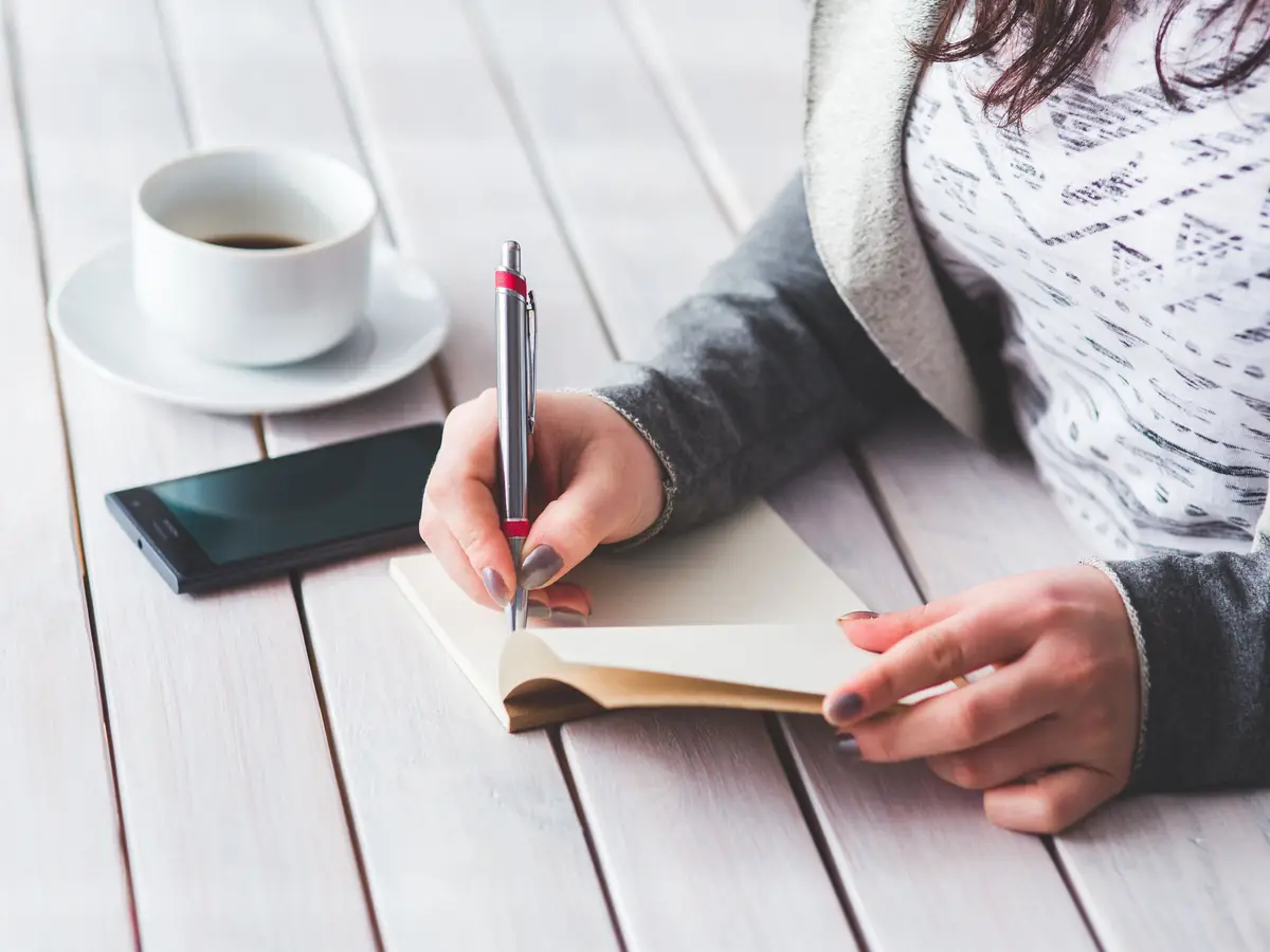 A hand writes in a small notebook on a wooden table beside a coffee cup and a phone.
