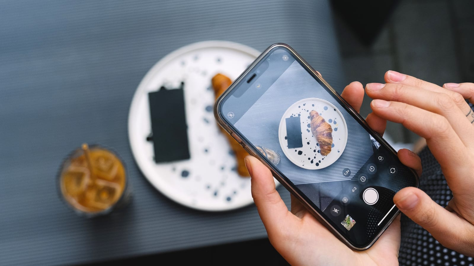Two hands frame a croissant on a phone screen at a cafe table, the shot a creator takes on a hosted visit.