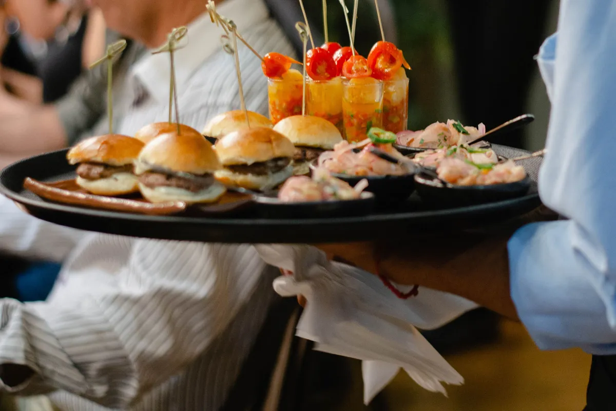 A server carries a round tray of sliders, small cups of salsa and bowls of seafood past seated guests.