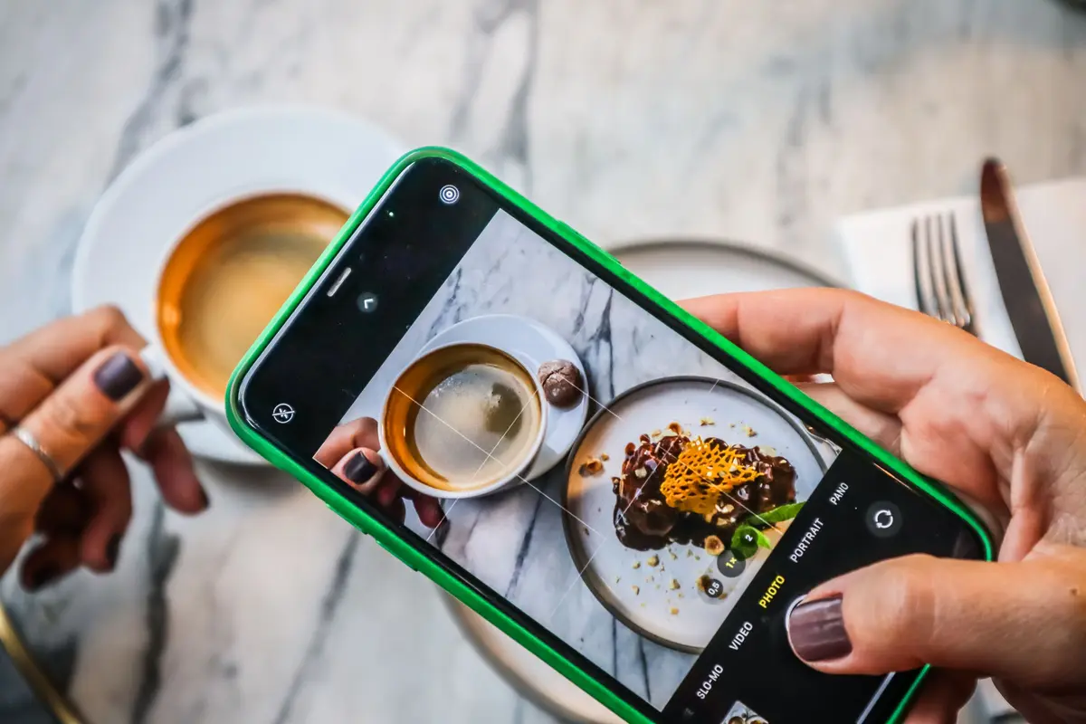 Hands frame a plated dessert and a cup of coffee on a marble table in a phone camera.