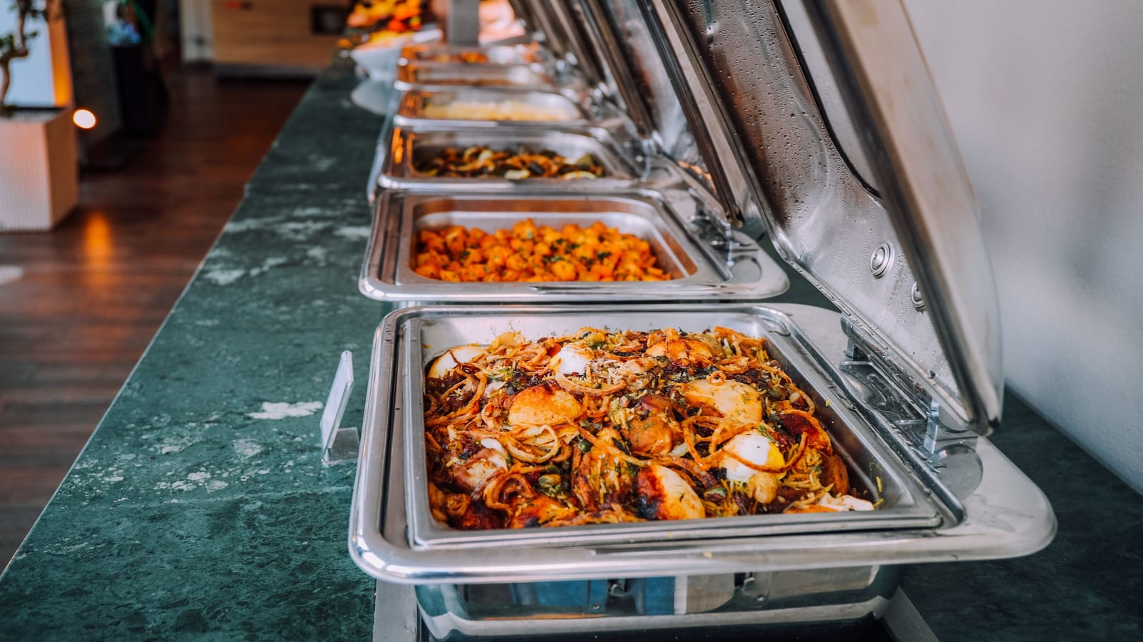 A row of stainless steel catering chafing dishes with their lids up on a long stone buffet counter, a rice dish topped with eggs and fried onions in the front pan.