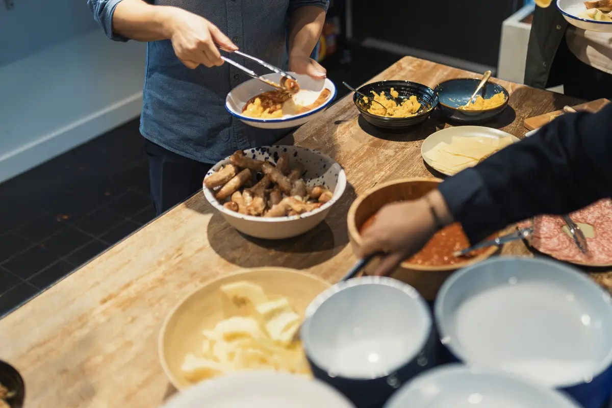 Guests at a workplace buffet serve themselves with tongs from bowls of sausages, scrambled eggs and beans, shown from the shoulders down.