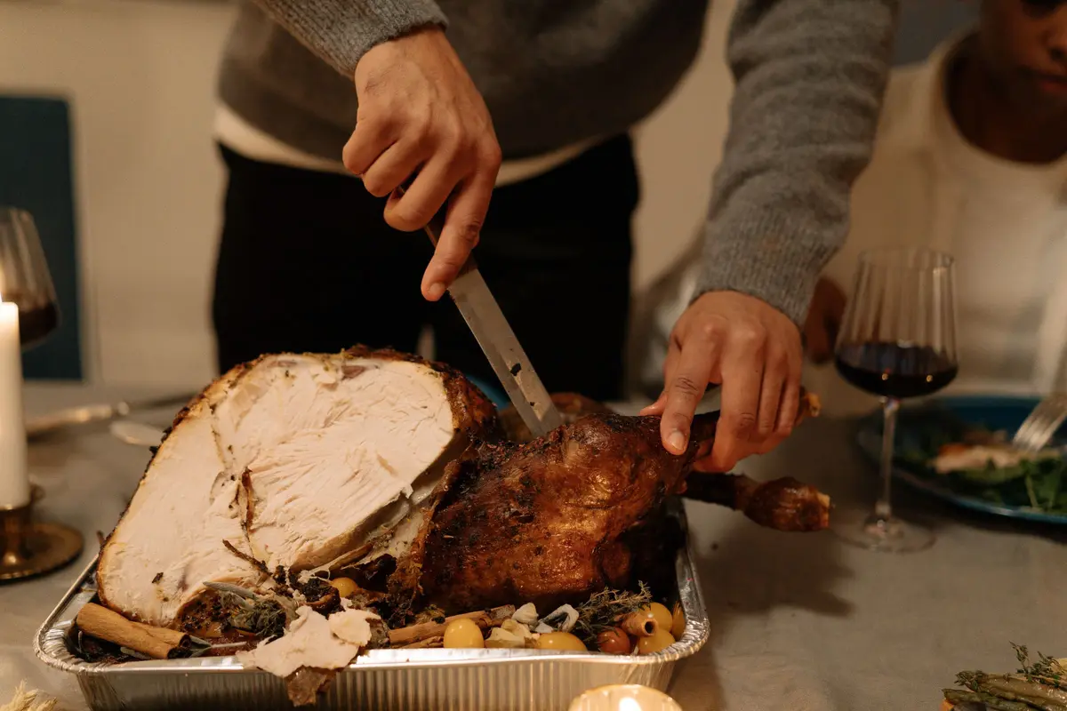 Hands carve slices of breast meat from a roast turkey sitting in a disposable foil pan.