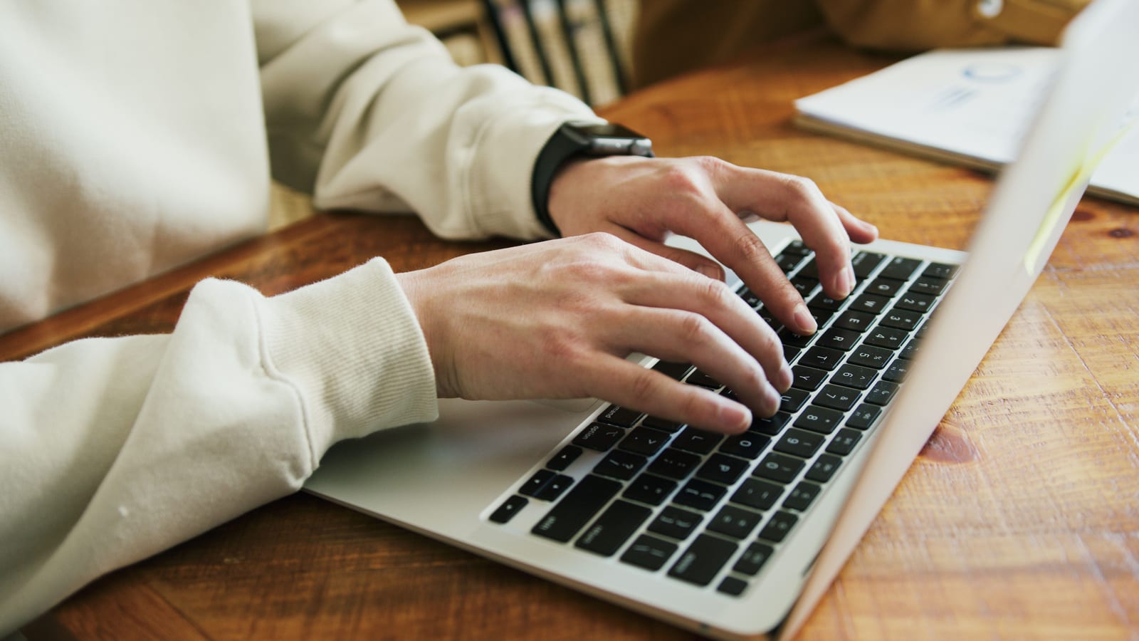 Two hands type on a laptop keyboard on a wooden desk.