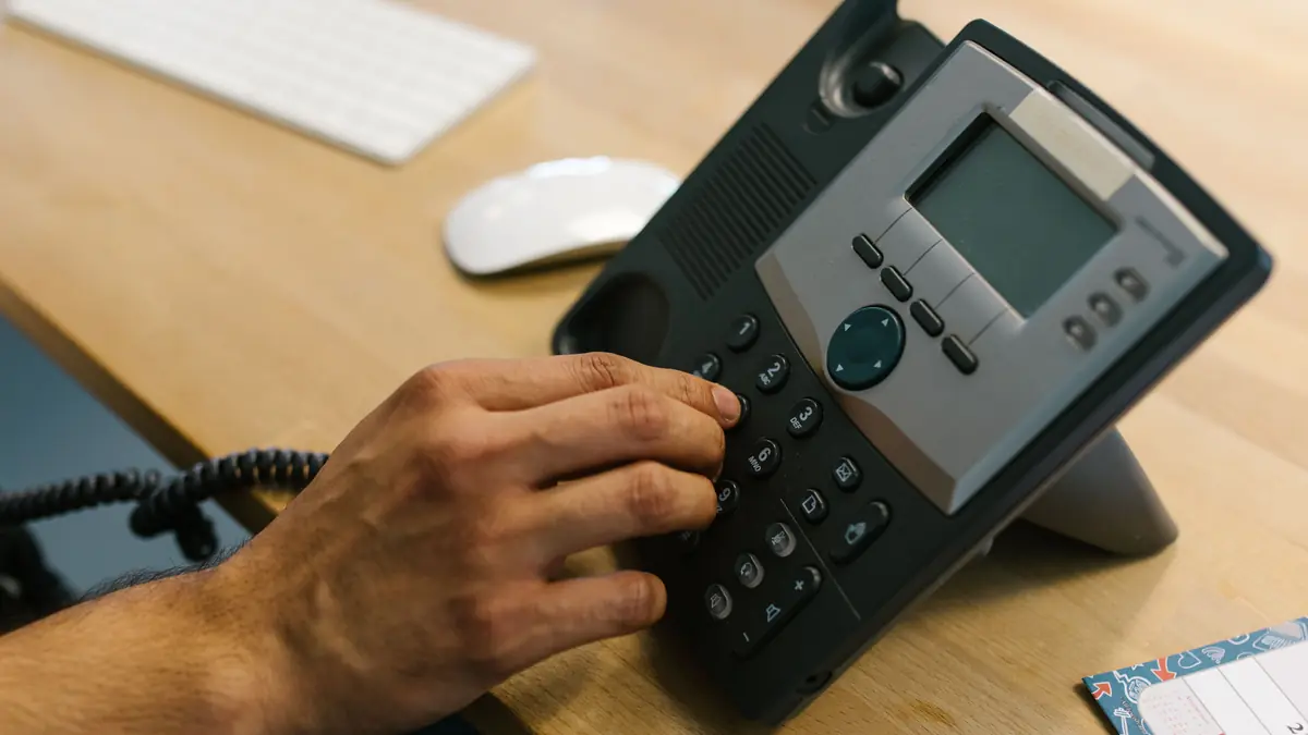 A hand presses the keys of a desk phone beside a keyboard and mouse.