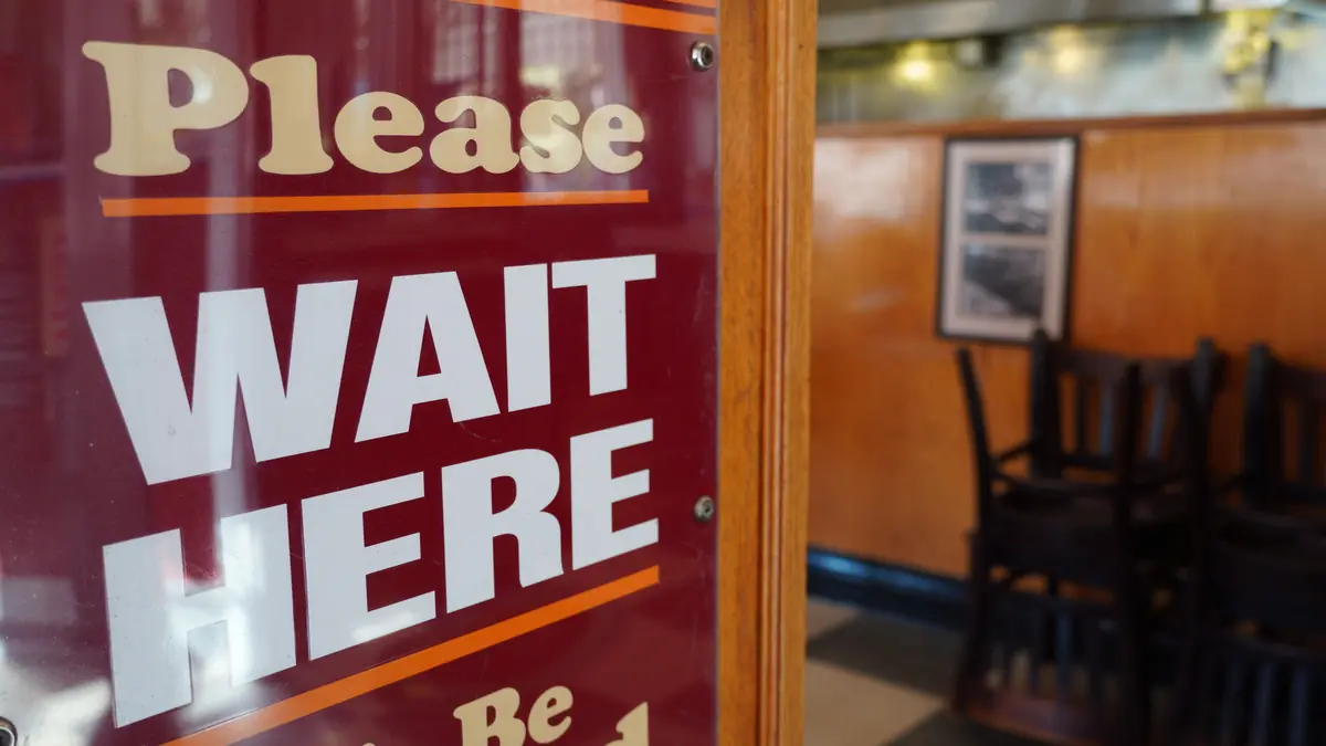 A red sign at a diner entrance reads “Please wait here”, with an empty dining room behind it.