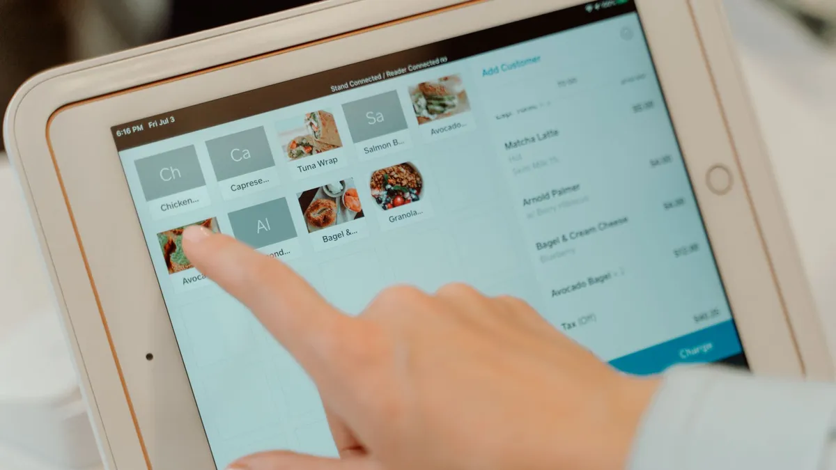 A finger taps a menu item tile on a tablet point-of-sale screen, with the open order listed on the right.