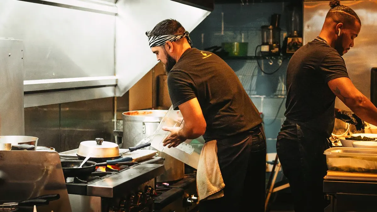 Two cooks work the range under a steel hood in a small kitchen, a pan on the flame and a stockpot behind it