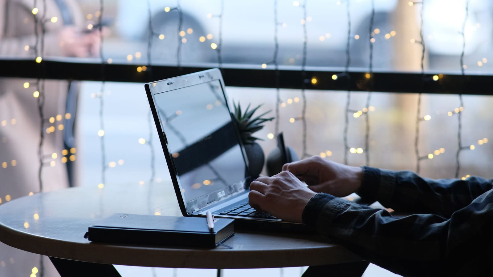 Hands typing on a laptop at a small round table by a window, string lights blurred outside, a notebook beside it.