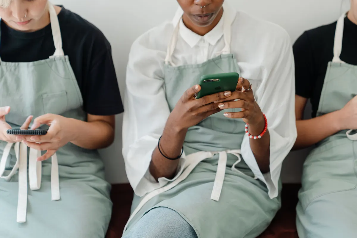 Three café staff in aprons check their phones behind the counter.