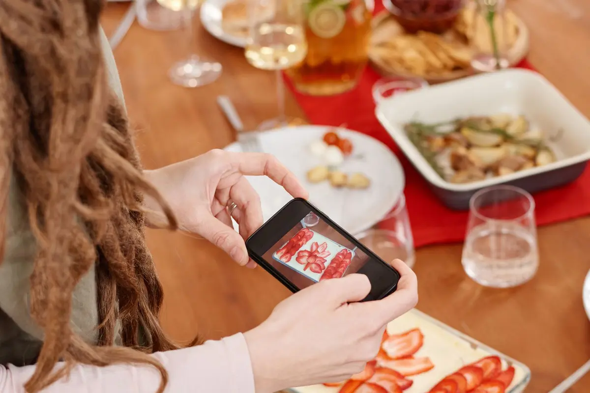 A guest’s hands hold a phone over a strawberry dessert on a set table, framing the shot before anyone takes a bite.