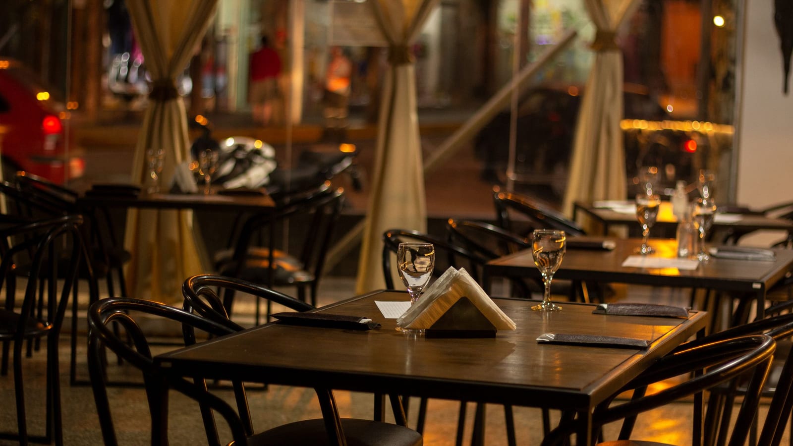 Set but empty tables in a small restaurant at night, with water glasses and a napkin holder on dark wood and the street blurred outside.