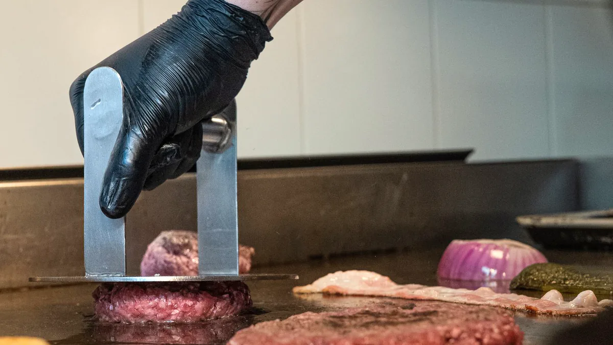 A cook in a black glove presses a burger patty flat on the griddle, more patties and a cut onion cooking beside it