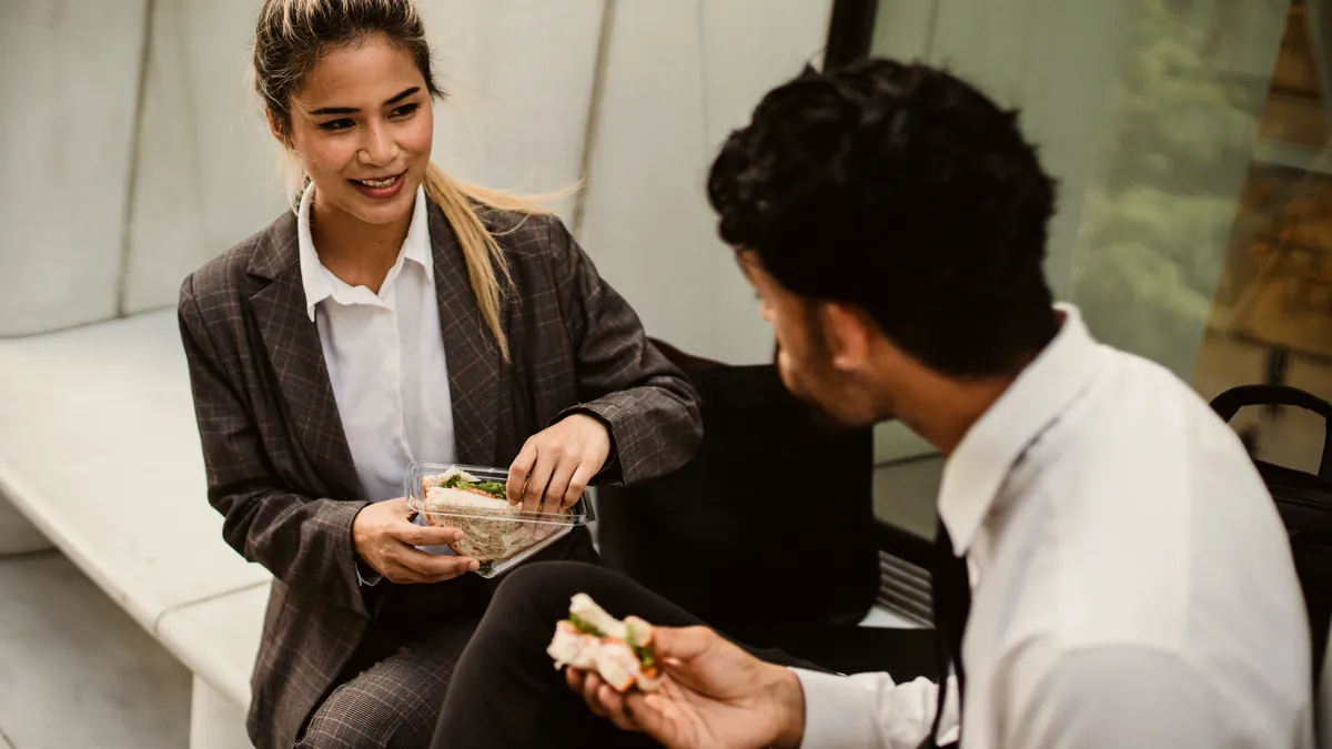 Two office workers, a woman in a grey blazer and a man in a white shirt, talk over sandwiches on a lunch break.